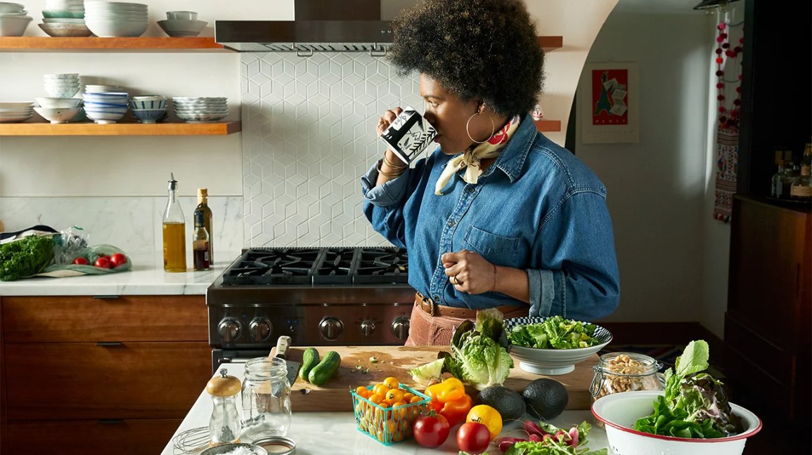 woman drinking from a mug and standing near a table containing tomatoes and other produce